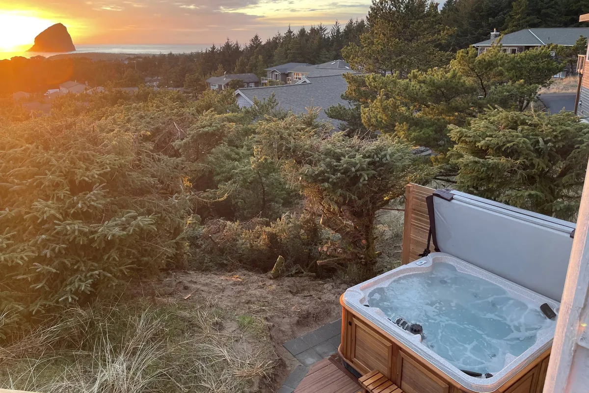 This image captures the hot tub on the deck at sunset, surrounded by trees and overlooking a distant rock formation. The scene exudes tranquility, with the warm glow of the sinking sun reflecting off the water, and the hot tub inviting guests to unwind while soaking in the natural beauty.