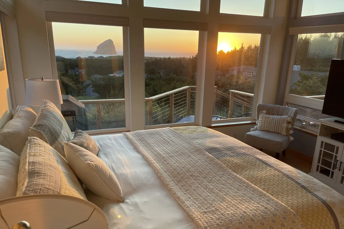 A serene master bedroom with a plush king-sized bed adorned with decorative pillows and a wall of large windows, offering a breathtaking sunset view over Haystack Rock. The wooden balcony railing outside enhances the sense of retreat and privacy.