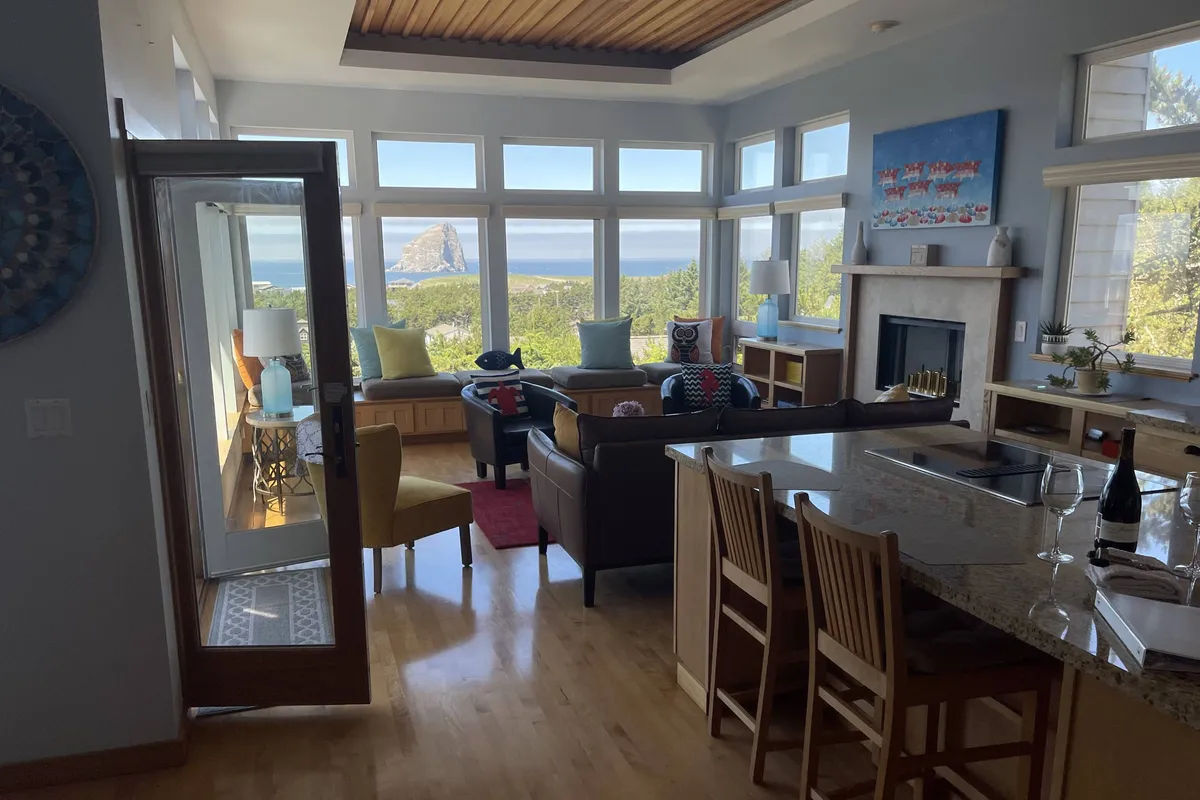 This image showcases an open-concept living area with large windows framing a sweeping ocean view, a cozy seating space, a kitchen island with bar stools, and vibrant local artwork on the walls. The scene is bathed in natural light, emphasizing the seamless blend of living, dining, and kitchen spaces.