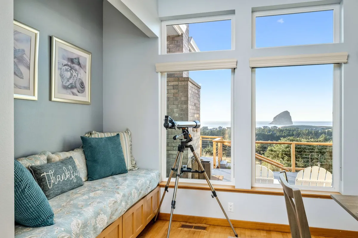 A tranquil living space with expansive windows framing the landscape, a telescope positioned for stargazing, and decorative pillows on a comfortable seating area. The view through the windows hints at the coast and open sky, creating a peaceful retreat for relaxation or inspiration.