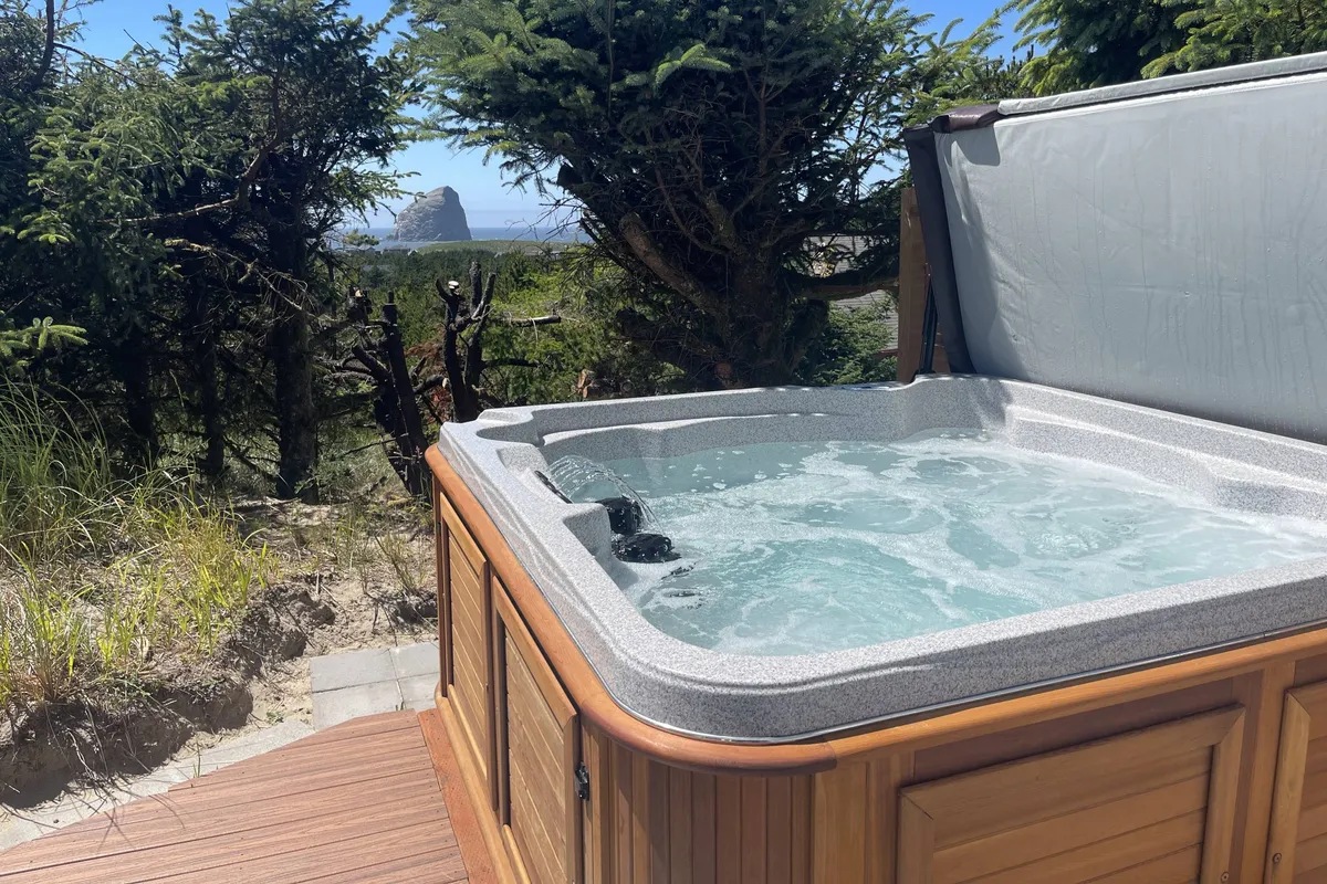An inviting exterior scene showing a hot tub surrounded by lush greenery, with a clear view of a distant rock formation (suggestive of Haystack Rock). The image hints at relaxation and the potential for wildlife watching or sunset viewing from the comfort of the property’s outdoor amenities.