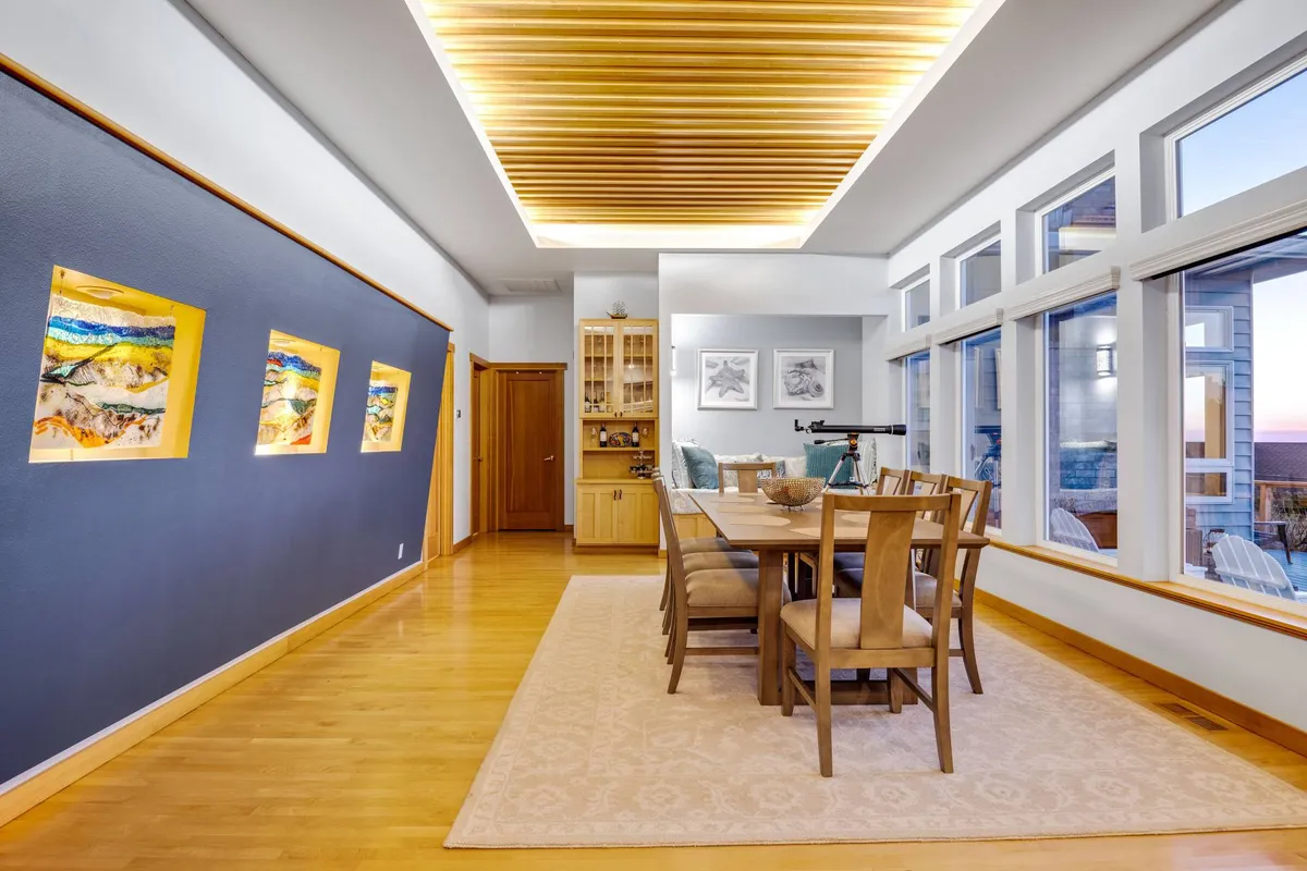 A dining area with a table set for family meals, backed by floor-to-ceiling windows that let in radiant light and offer a glimpse of the ocean. Local artwork adorns the wall, and plush seating invites guests to gather for meals or conversation.