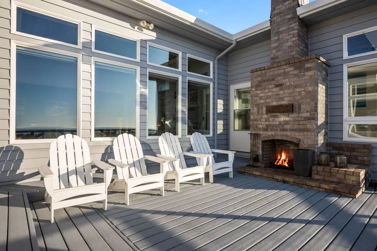 A cozy exterior shot of the deck, featuring Adirondack chairs positioned around a brick fireplace. The large windows of the house reflect the evening light, and the setup encourages conversation, warmth, and togetherness under the stars.