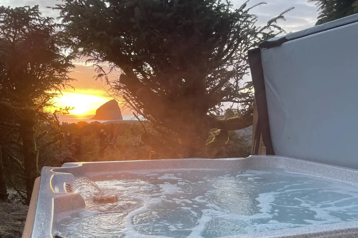 An inviting exterior shot featuring a jacuzzi nestled among surrounding trees, set against a glowing sunset sky. The image captures the spa's proximity to nature and hints at both woodland and coastal elements beyond.