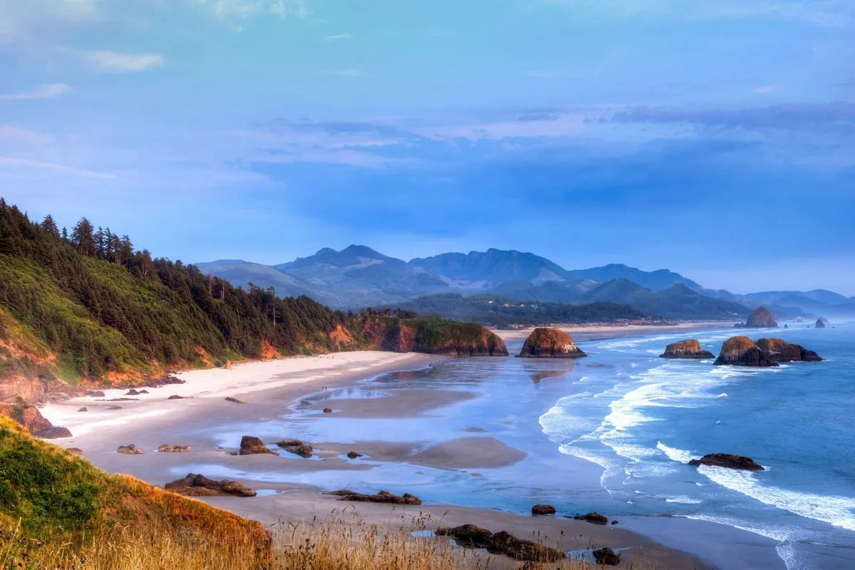 A sweeping panoramic view showcasing dramatic rock formations (likely Haystack Rock), ocean waves, a broad sandy beach, and the surrounding natural landscape, including forest and mountains. The lighting suggests either sunrise or sunset, enhancing the visual drama and serenity. This image evokes the wild, memorable beauty of the Oregon coast.