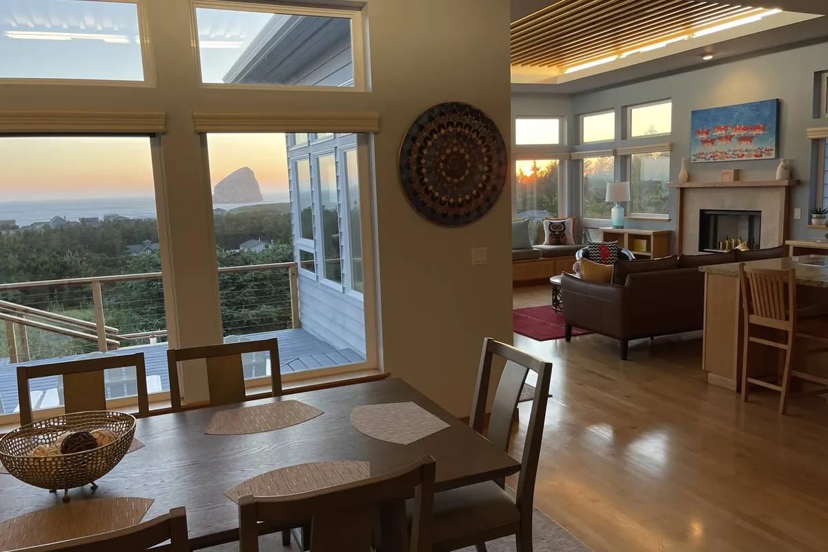 A dining table set near expansive windows with ocean views, a cozy seating area with a fireplace, and a decorative bowl as a centerpiece. The space is filled with natural light, offering a tranquil setting for family meals or peaceful reflection as the sun sets.