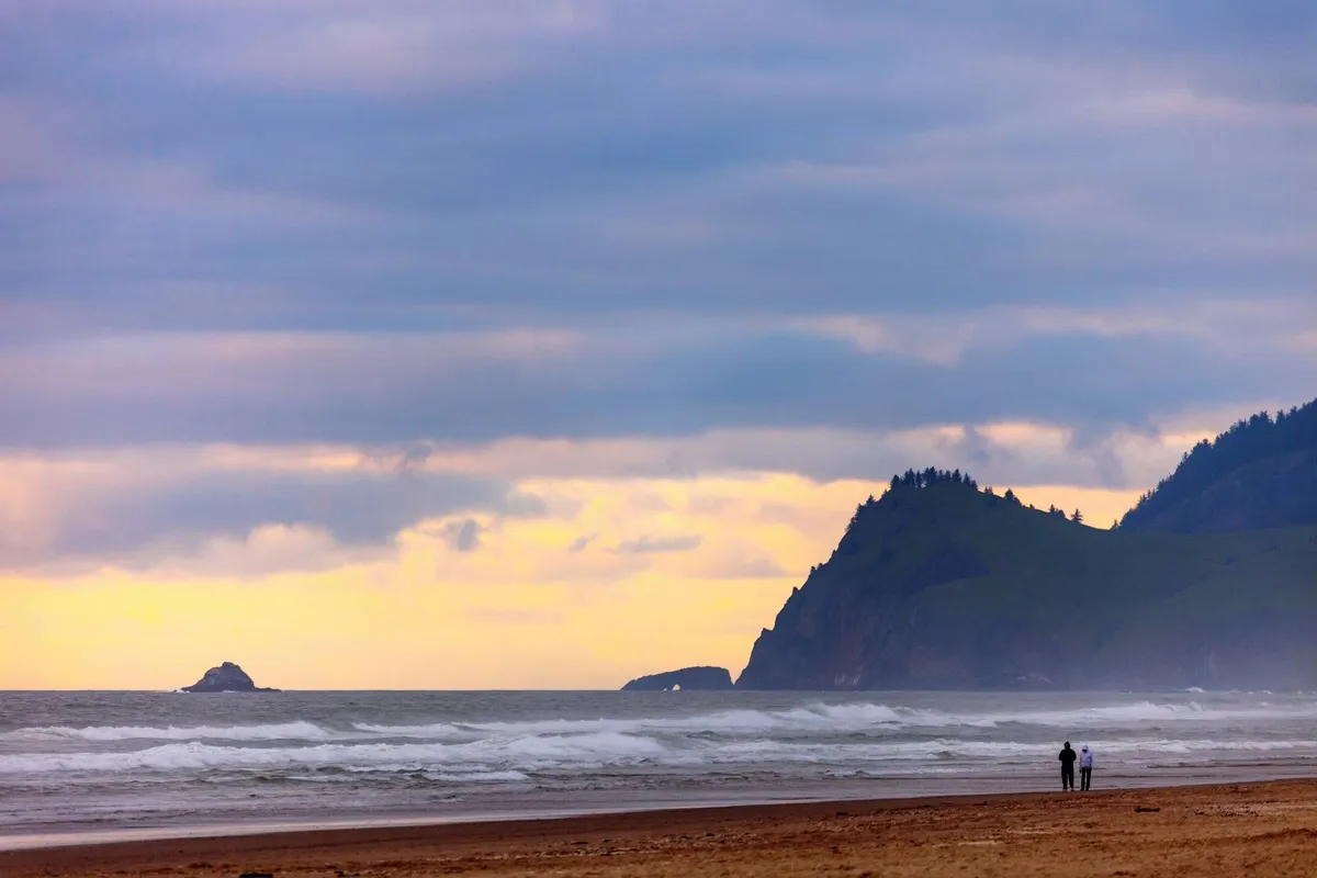 An exterior view featuring the broad, sandy expanse of Pacific City Beach with ocean waves rolling in, distant cliffs, and a couple walking along the shoreline. The late-day light casts a warm glow, evoking the feeling of sunset. The scene captures both the iconic Oregon coast landscape and the leisurely activities that families and couples enjoy, such as beachcombing and strolling.