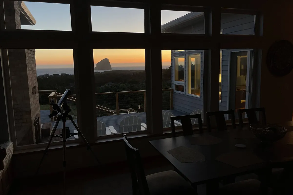 An inviting indoor space featuring a dining table, adirondack chairs, a telescope, and expansive windows framing a scenic sunset view. The space exudes tranquility and offers opportunities for stargazing, sunset watching, and intimate meals.