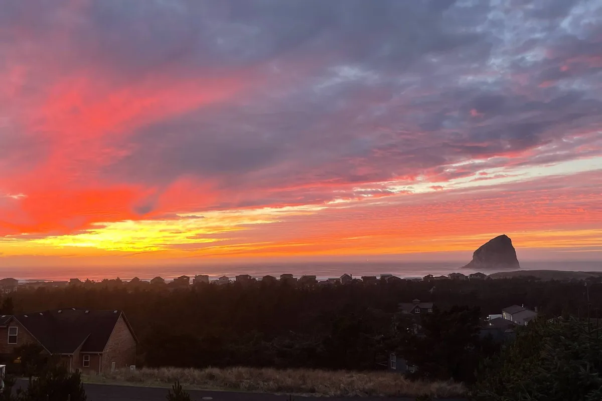 A breathtaking view of a vibrant sunset over Haystack Rock, with silhouetted houses in the foreground. The scene captures the serene atmosphere and natural beauty that guests can experience while relaxing outdoors at the property.