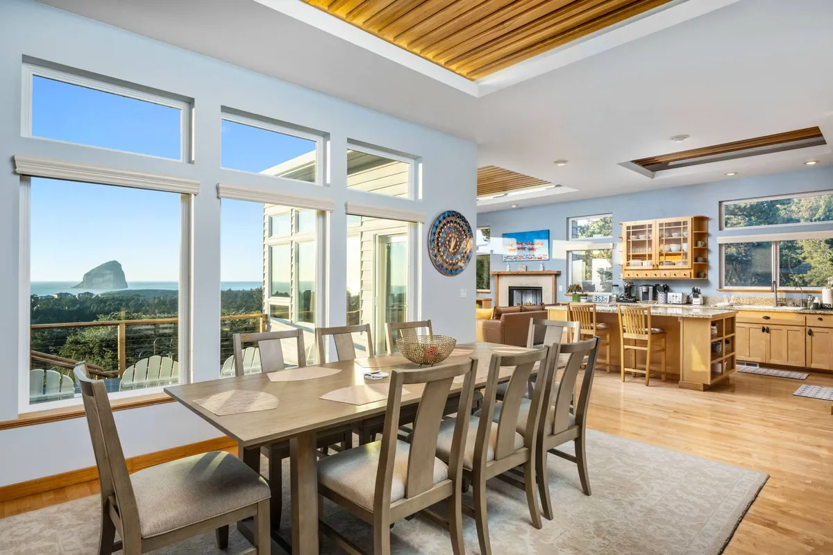 The image showcases a bright, open-concept dining and kitchen area with a spacious table set for a gathering. Floor-to-ceiling windows frame breathtaking ocean views, capturing the golden glow of sunset over the Pacific. The scene is warm and inviting, ideal for family meals and holiday celebrations while overlooking the sea.