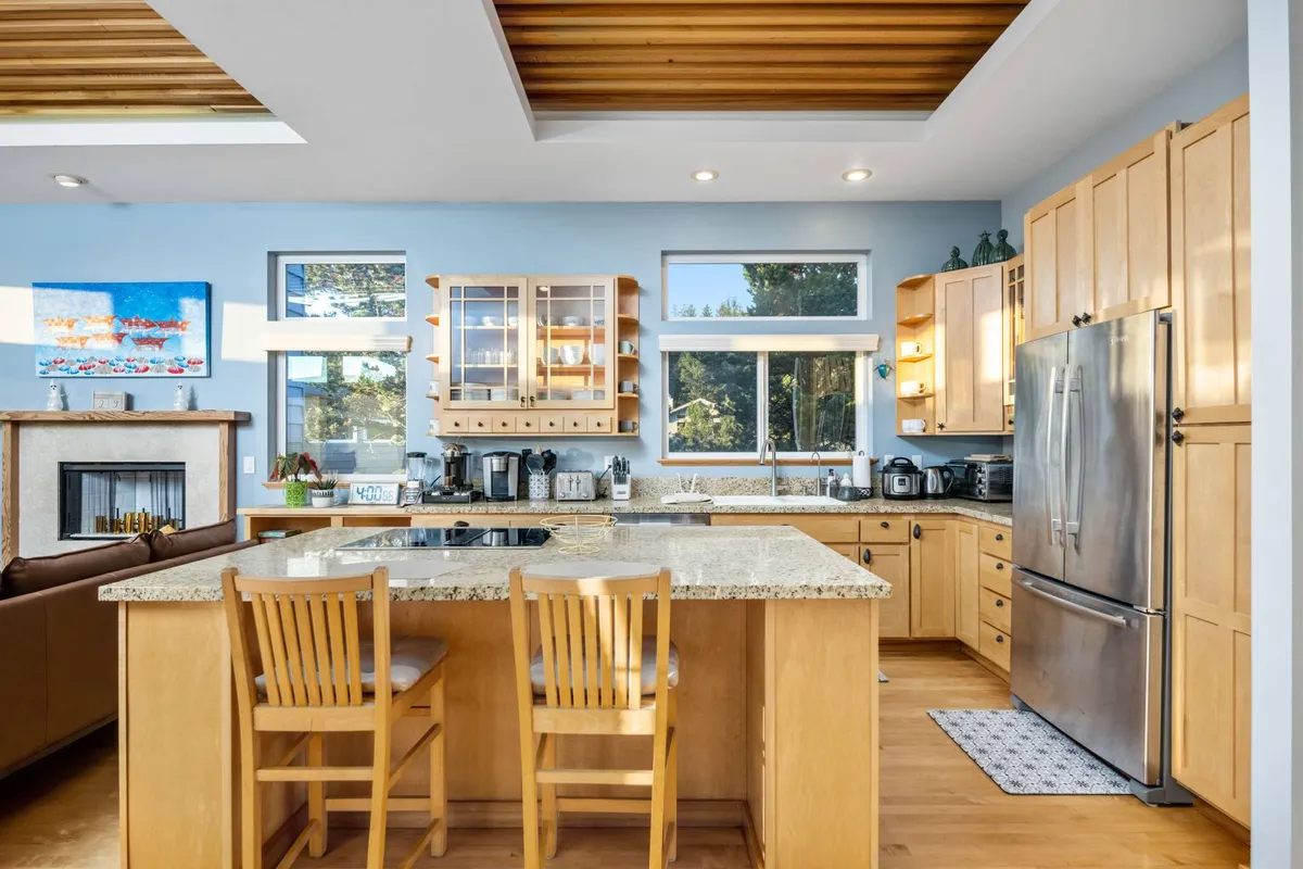 A chef’s kitchen featuring a spacious central island with seating, granite countertops, natural wood cabinetry, modern appliances, and large windows that introduce abundant daylight and frame scenic views. The layout invites both culinary creativity and social interaction.